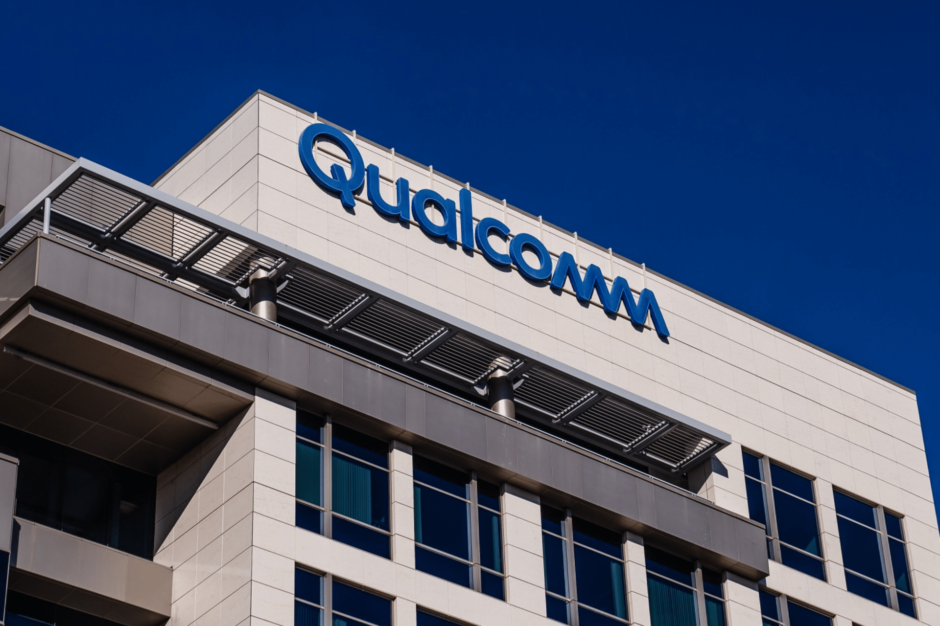 A modern office building with the Qualcomm logo displayed near the roof against a clear blue sky. Known for its role in smartphone prices and court battles, the structure features large windows and contemporary architectural elements.