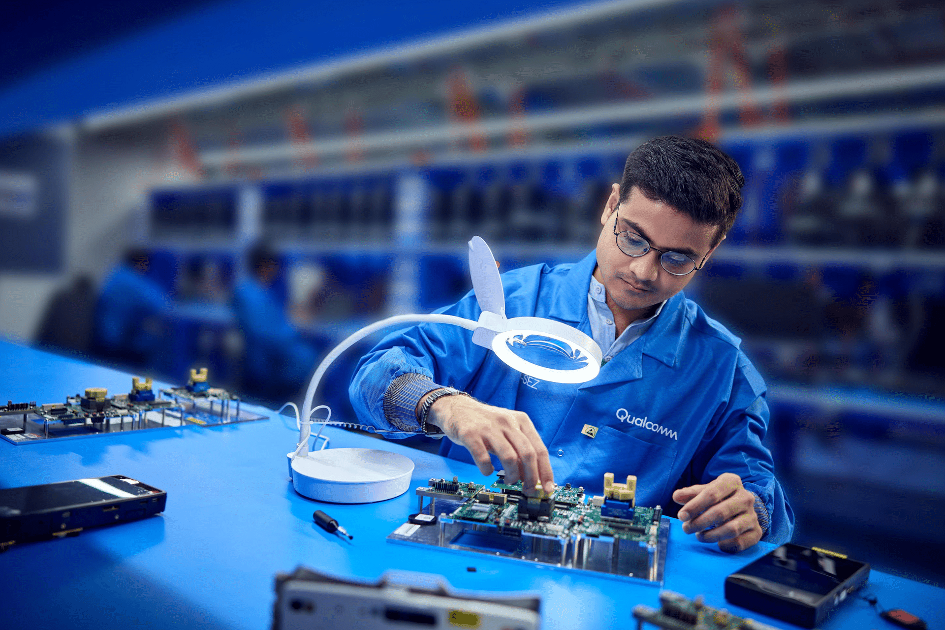 A man in a blue lab coat works on electronic circuit boards at a blue workstation, using tools and a magnifying lamp—his meticulous work reflects the ongoing Qualcomm court battle over smartphone prices, with shelves of components blurred in the background.