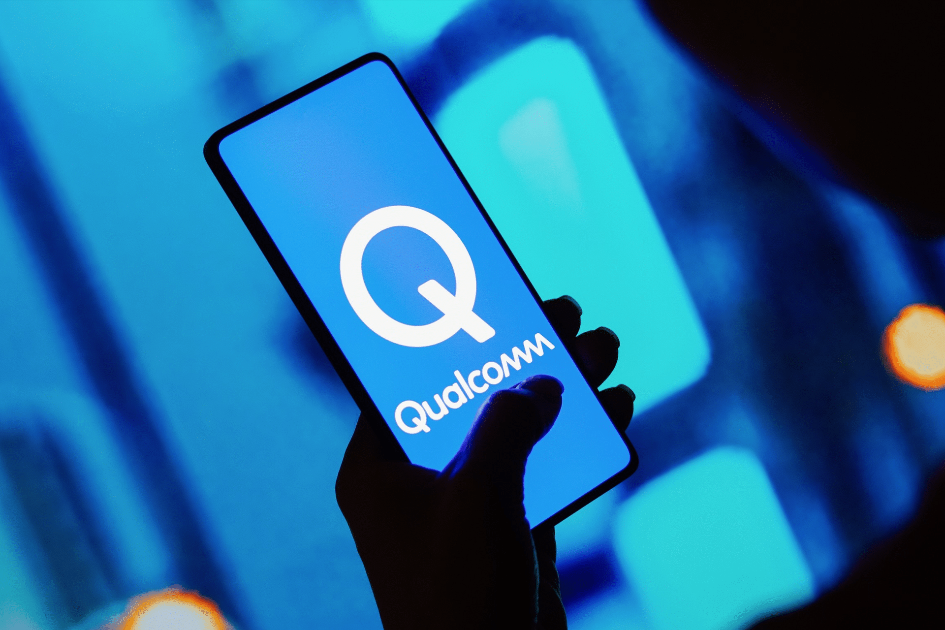 A person holds a smartphone displaying the Qualcomm logo on its screen, with a blurred blue background and some orange light spots behind—a subtle nod to the ongoing Qualcomm court battle over smartphone prices.