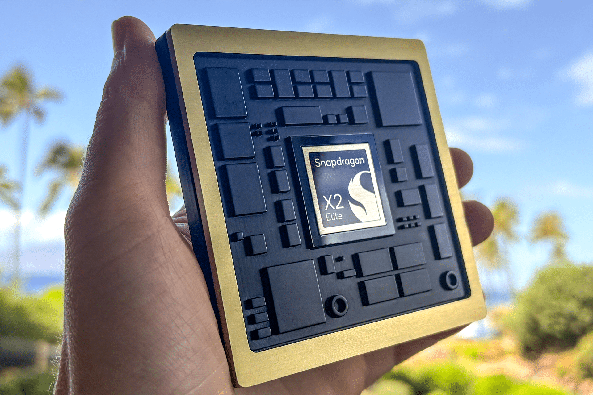 A hand holds a large display model of a Snapdragon X Elite processor chip—one of Qualcomm AI chips—outdoors, with palm trees and a blue sky blurred in the background.
