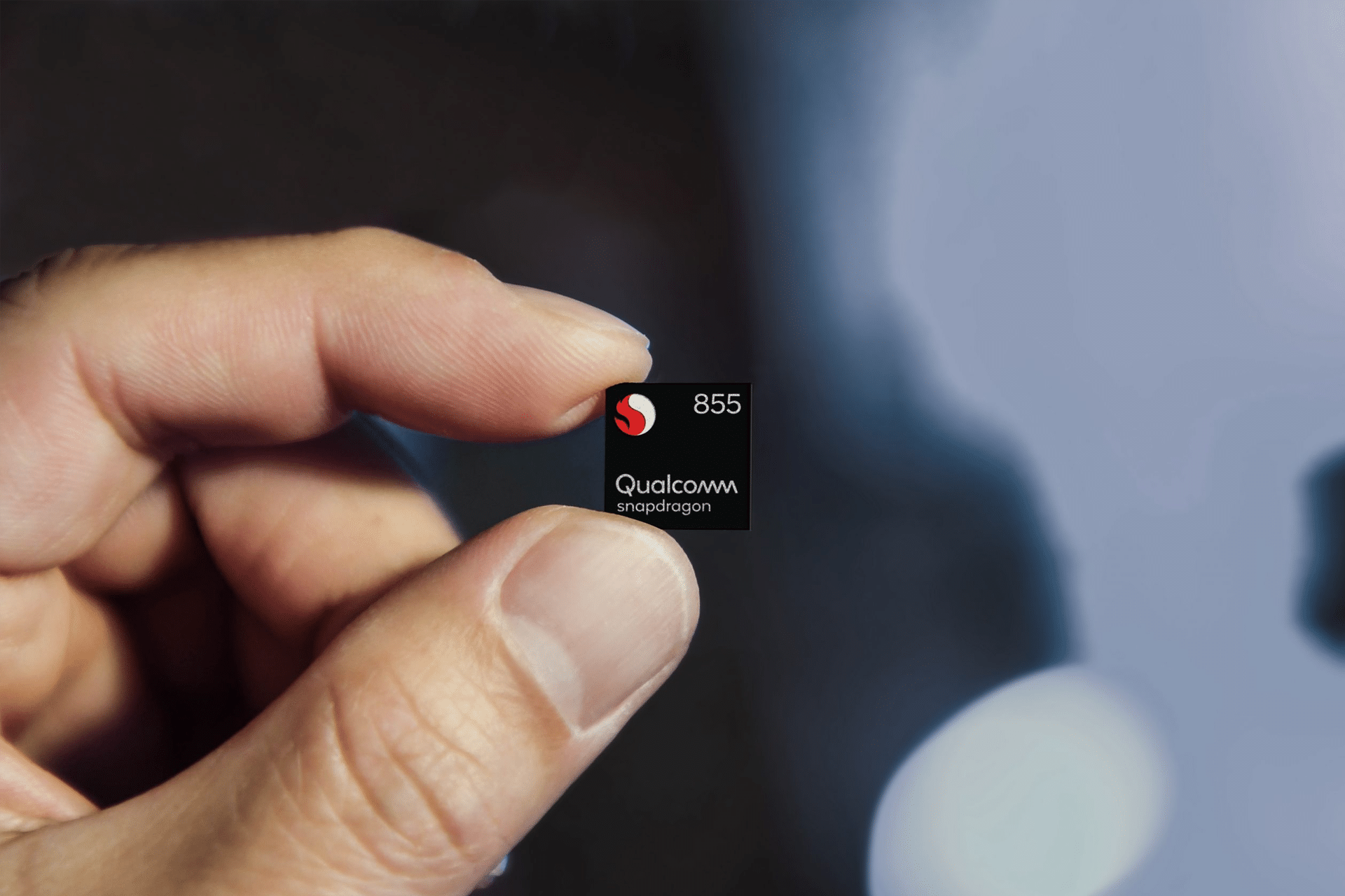 A hand holds a small black Qualcomm Snapdragon 855 chip, featuring the logo and name, against a blurred blue and black background—showcasing the innovation behind Qualcomm AI chips.