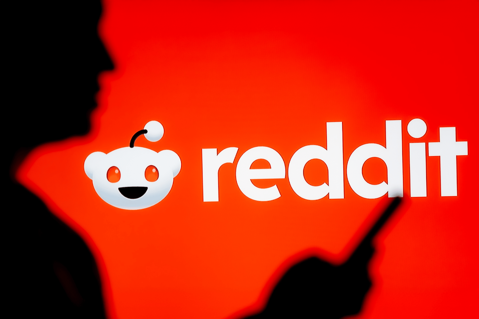 A silhouette of a person holding a smartphone stands before a bright red background featuring the Reddit logo and text, subtly referencing recent Reddit AI job cuts.