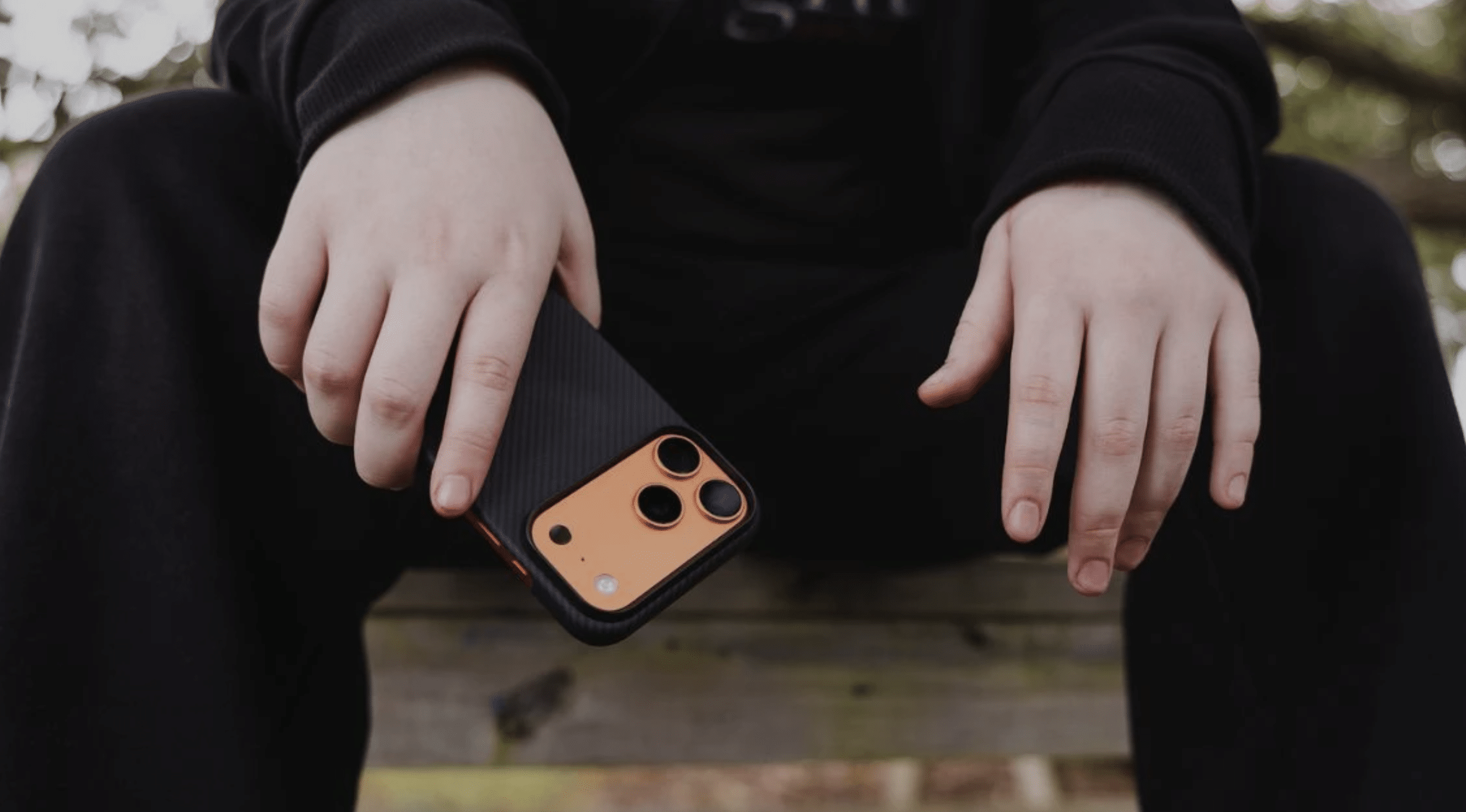 A person sitting on a bench holds a smartphone with a thin iPhone 17 Pro Max case in orange and black, their hands resting on their knees. The person's face is not visible.