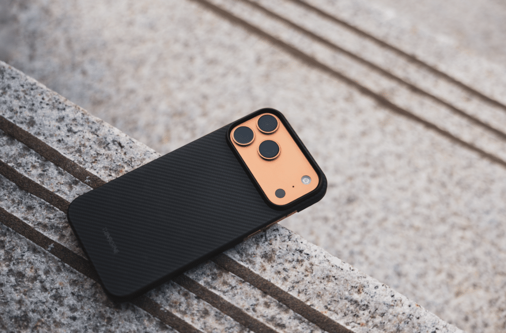 A thin iPhone 17 Pro Max case with a black carbon fiber finish and an orange camera module rests on a textured stone surface with parallel lines.