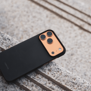 A thin iPhone 17 Pro Max case with a black carbon fiber finish and an orange camera module rests on a textured stone surface with parallel lines.