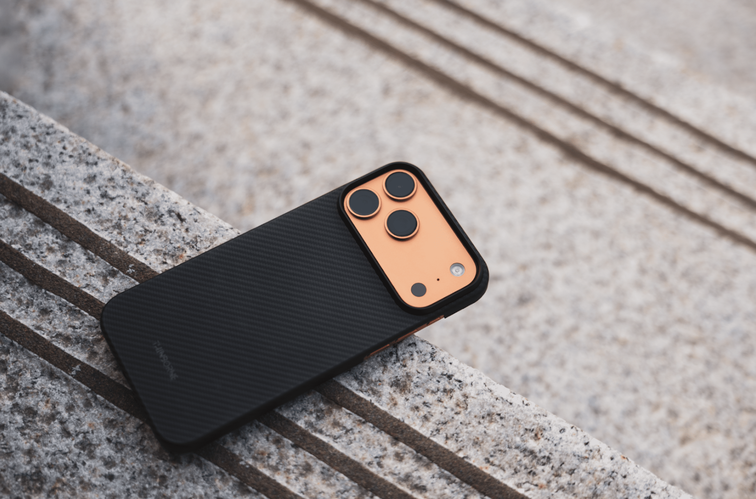 A thin iPhone 17 Pro Max case with a black carbon fiber finish and an orange camera module rests on a textured stone surface with parallel lines.