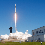 A SpaceX rocket launches into a clear blue sky, leaving a bright trail of flames and smoke, as speculation grows around the potential Globalstar SpaceX acquisition; the SpaceX building stands visible in the foreground.
