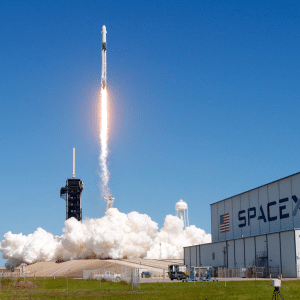A SpaceX rocket launches into a clear blue sky, leaving a bright trail of flames and smoke, as speculation grows around the potential Globalstar SpaceX acquisition; the SpaceX building stands visible in the foreground.