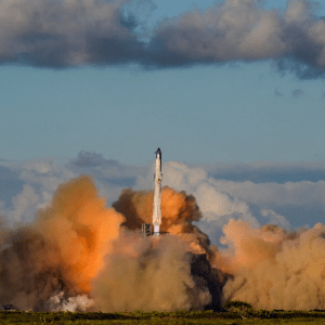 A rocket from the SpaceX 11th Starship test flight launches into the sky, surrounded by thick clouds of orange and gray smoke, with a backdrop of blue sky and scattered clouds above a green landscape.