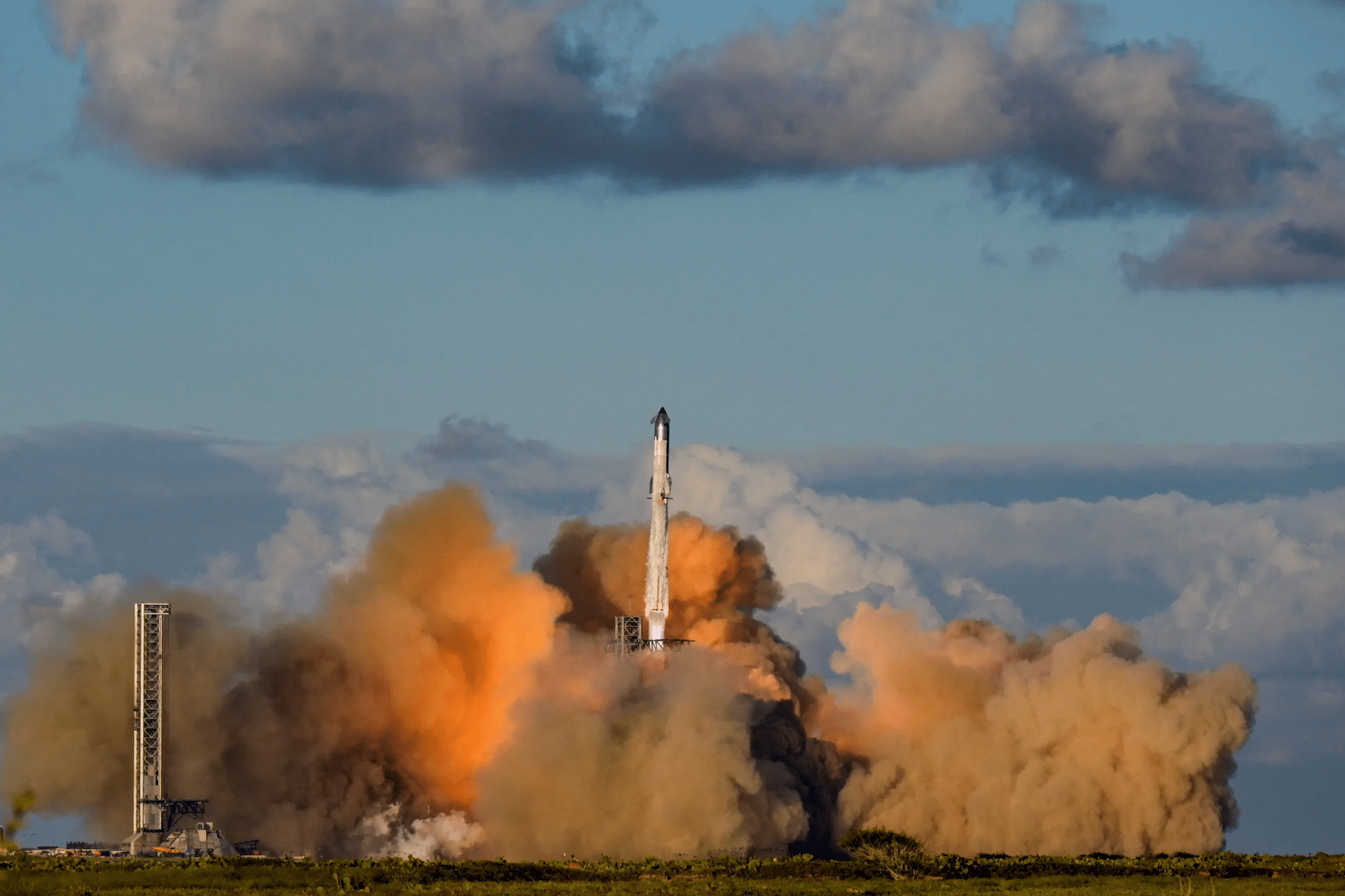 A rocket from the SpaceX 11th Starship test flight launches into the sky, surrounded by thick clouds of orange and gray smoke, with a backdrop of blue sky and scattered clouds above a green landscape.