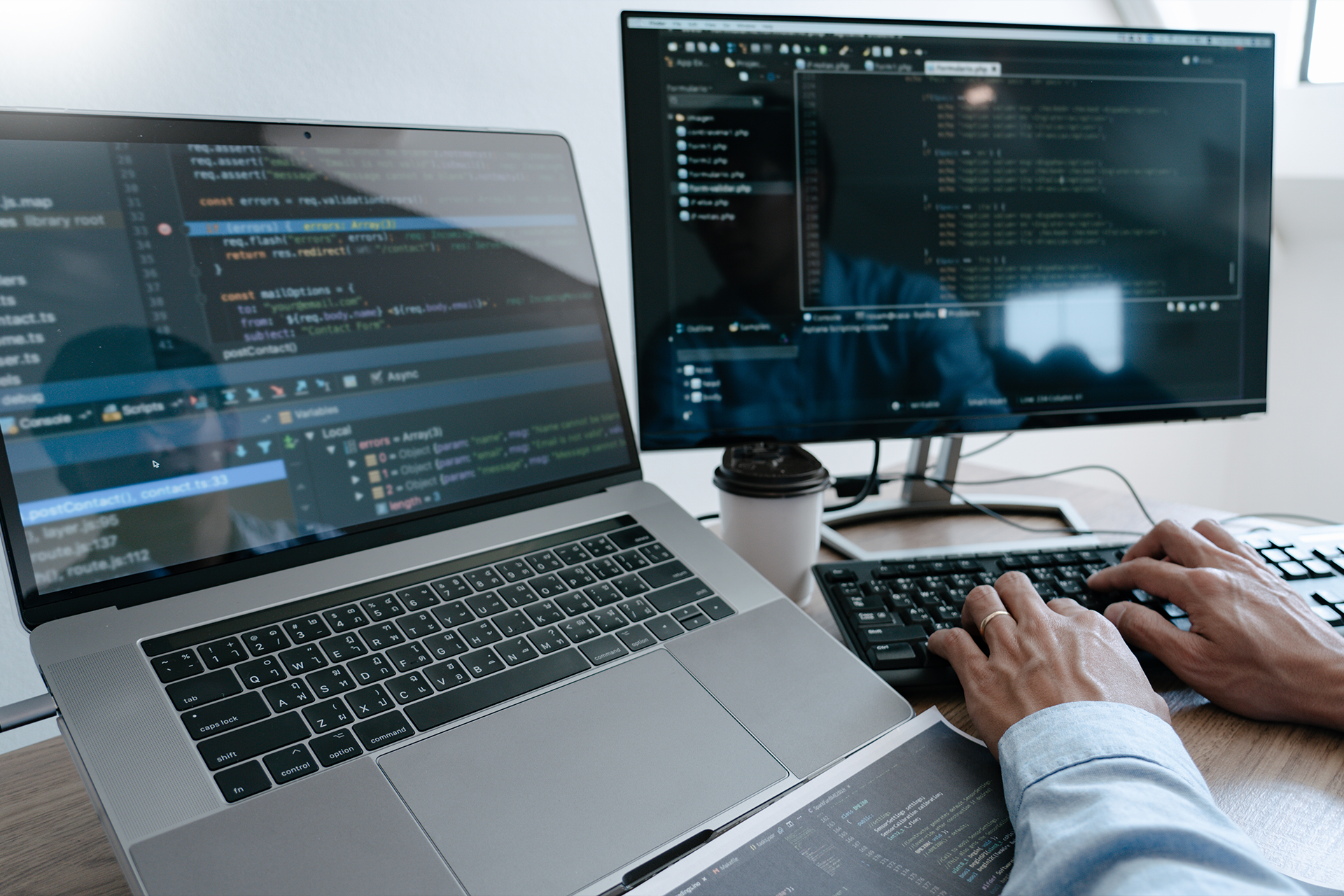 A person types on a keyboard at a desk with a laptop and monitor displaying code, illustrating the day-to-day work in software development—a common scene for computer science graduates entering the job market. A takeaway coffee cup sits nearby.
