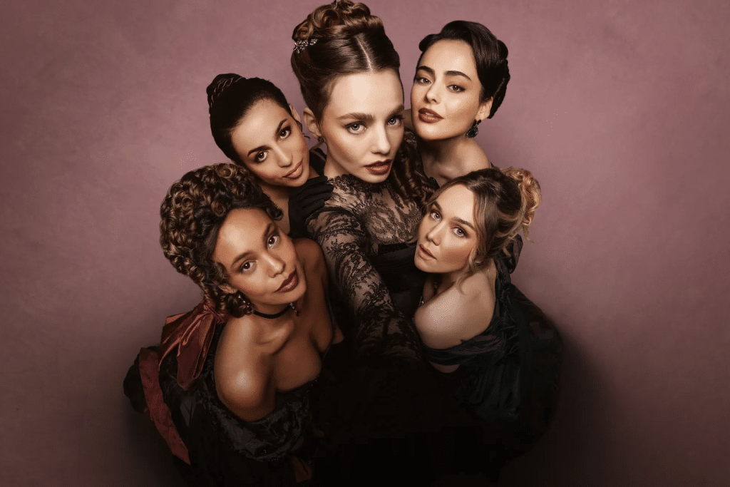 Five women in elegant, period-style dresses pose closely together, looking up at the camera against a pink background. Their confident, intense expressions and elaborate hairstyles evoke the bold spirit of The Buccaneers season three.