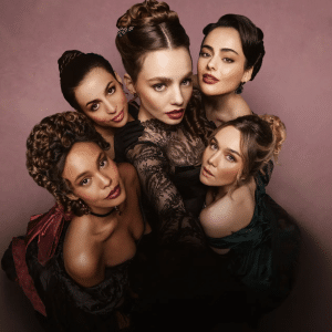 Five women in elegant, period-style dresses pose closely together, looking up at the camera against a pink background. Their confident, intense expressions and elaborate hairstyles evoke the bold spirit of The Buccaneers season three.