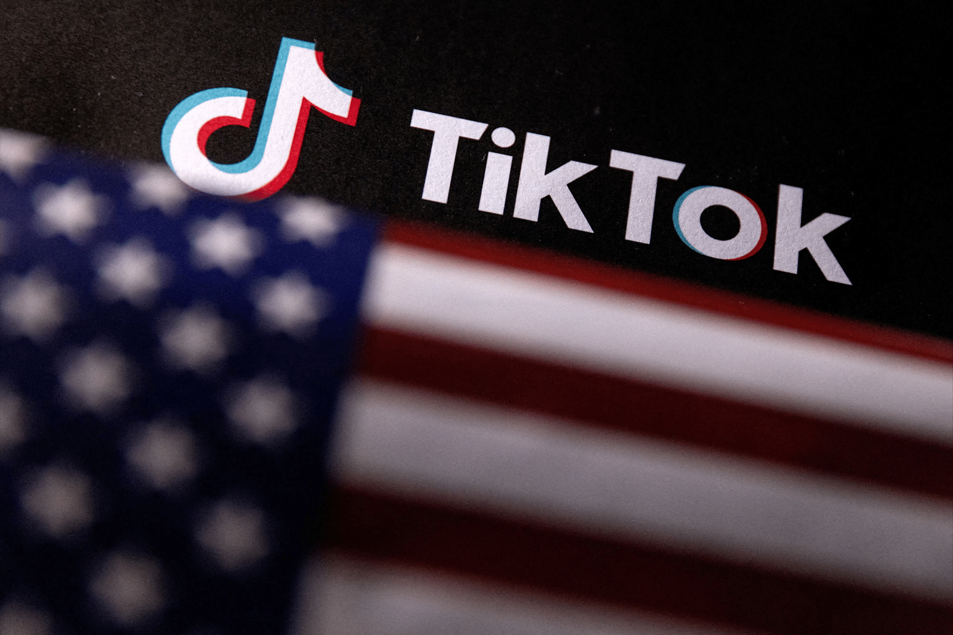 The TikTok logo and name are displayed on a black background, with a blurred American flag in the foreground—hinting at ongoing TikTok deal tensions between the U.S. and China.