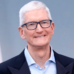 A man with short white hair and glasses, wearing a light blue collared shirt and a dark jacket, smiles while standing in front of a plain, light blue-gray background, embodying the calm confidence of Tim Cook Apple leadership.