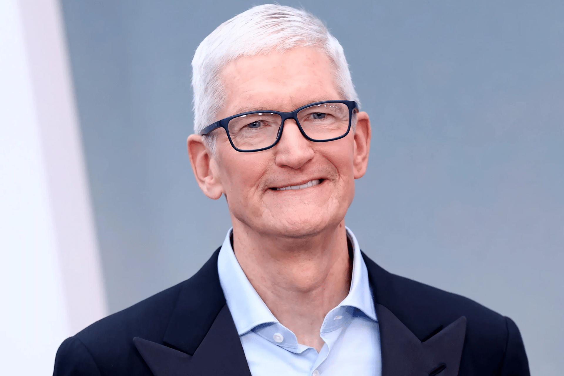 A man with short white hair and glasses, wearing a light blue collared shirt and a dark jacket, smiles while standing in front of a plain, light blue-gray background, embodying the calm confidence of Tim Cook Apple leadership.