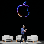Tim Cook walks on stage in front of three gray armchairs, with a large, glowing Apple logo above him, ready to introduce Apple Intelligence against a striking black background.