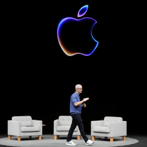 Tim Cook walks on stage in front of three gray armchairs, with a large, glowing Apple logo above him, ready to introduce Apple Intelligence against a striking black background.