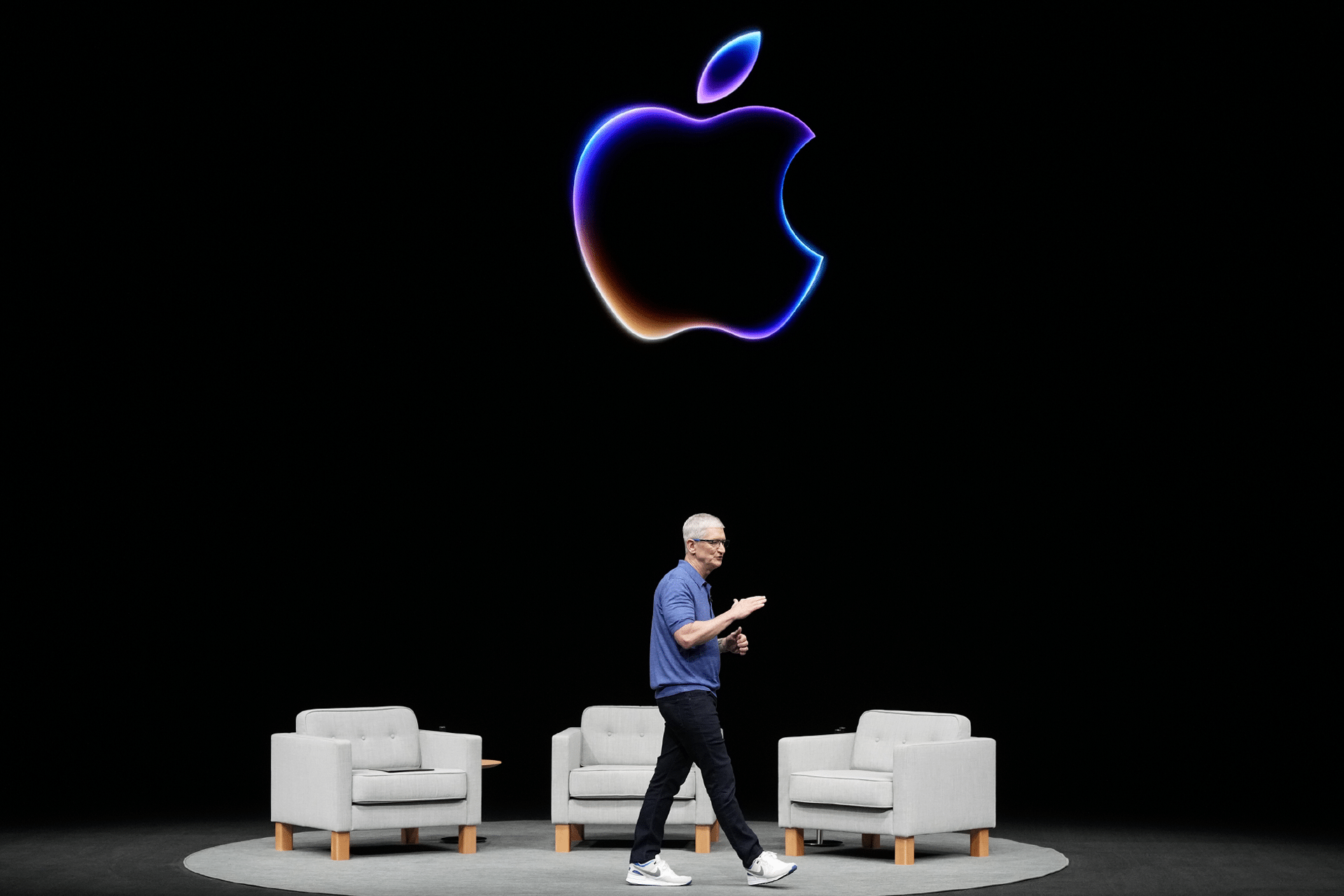 Tim Cook walks on stage in front of three gray armchairs, with a large, glowing Apple logo above him, ready to introduce Apple Intelligence against a striking black background.