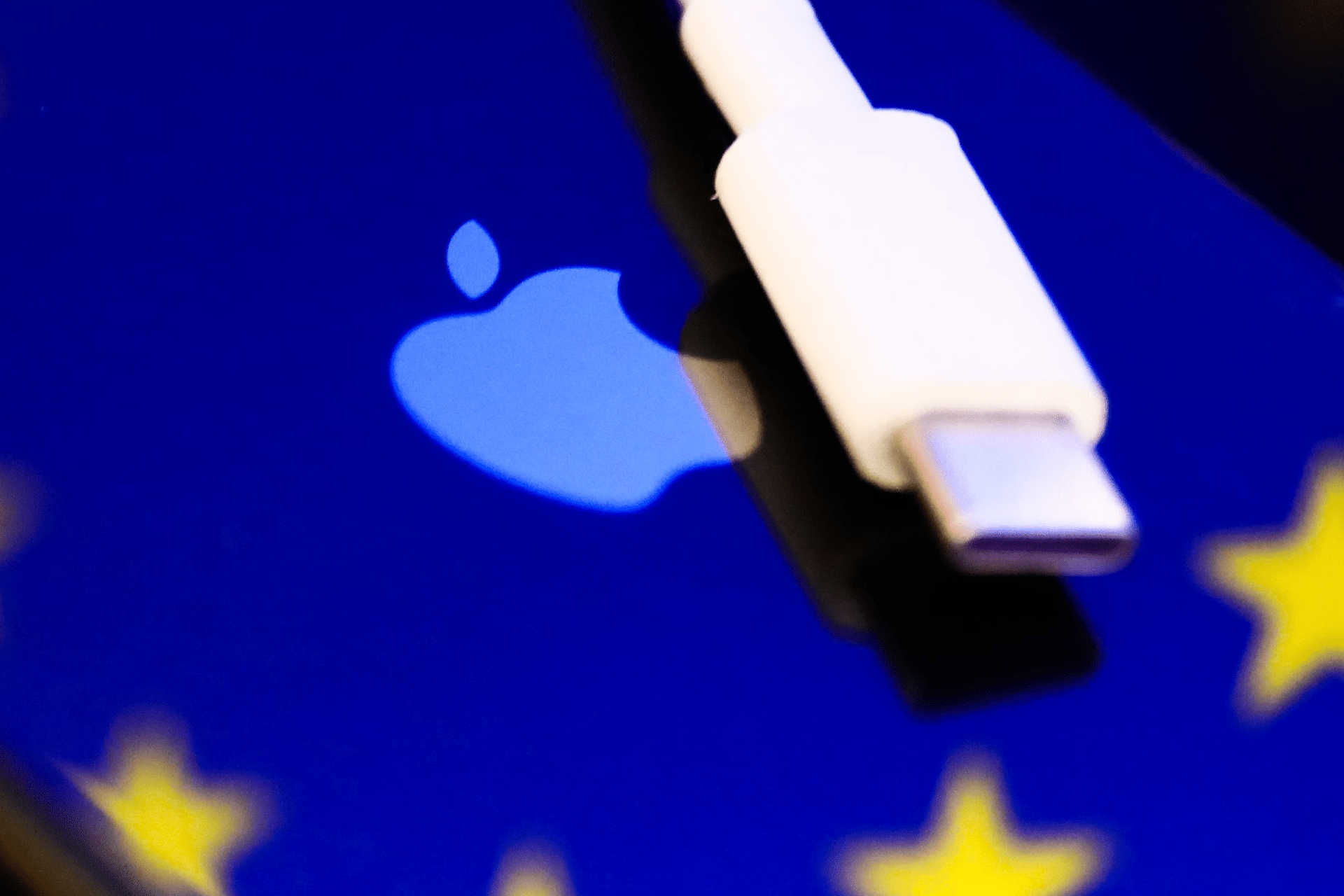 A white USB-C connector rests on a blue surface displaying the Apple logo and part of yellow stars, subtly evoking the ongoing Apple EU dispute over charging standards and regulations.