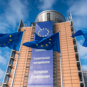 Three European Union flags wave in front of the European Commission headquarters in Brussels, where recent headlines have focused on the Apple EU dispute, highlighted by the modern building’s banner displaying the EU emblem and "European Commission" in many languages.