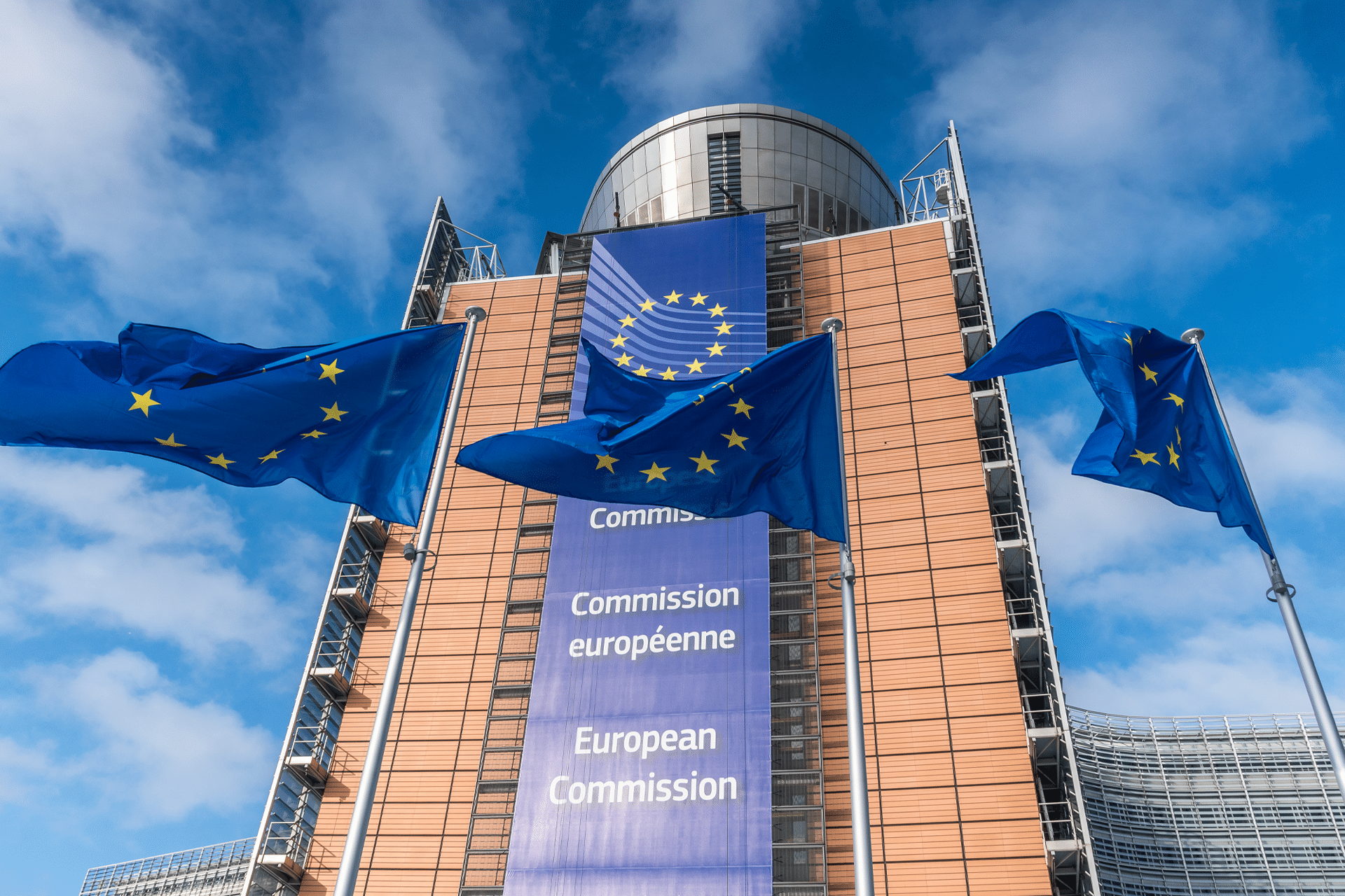 Three European Union flags wave in front of the European Commission headquarters in Brussels, where recent headlines have focused on the Apple EU dispute, highlighted by the modern building’s banner displaying the EU emblem and "European Commission" in many languages.