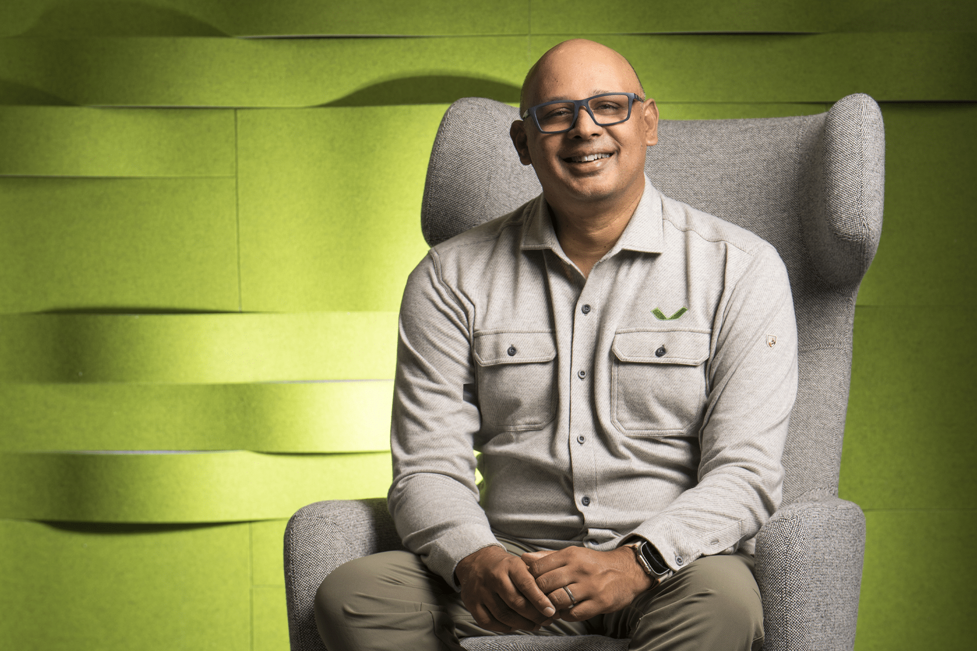 A smiling, bald man with glasses sits in a gray armchair. He wears a light gray shirt with chest pockets, khaki pants, and a smartwatch. The background’s textured green panels subtly reference new beginnings like Veeam acquires Securiti.ai.