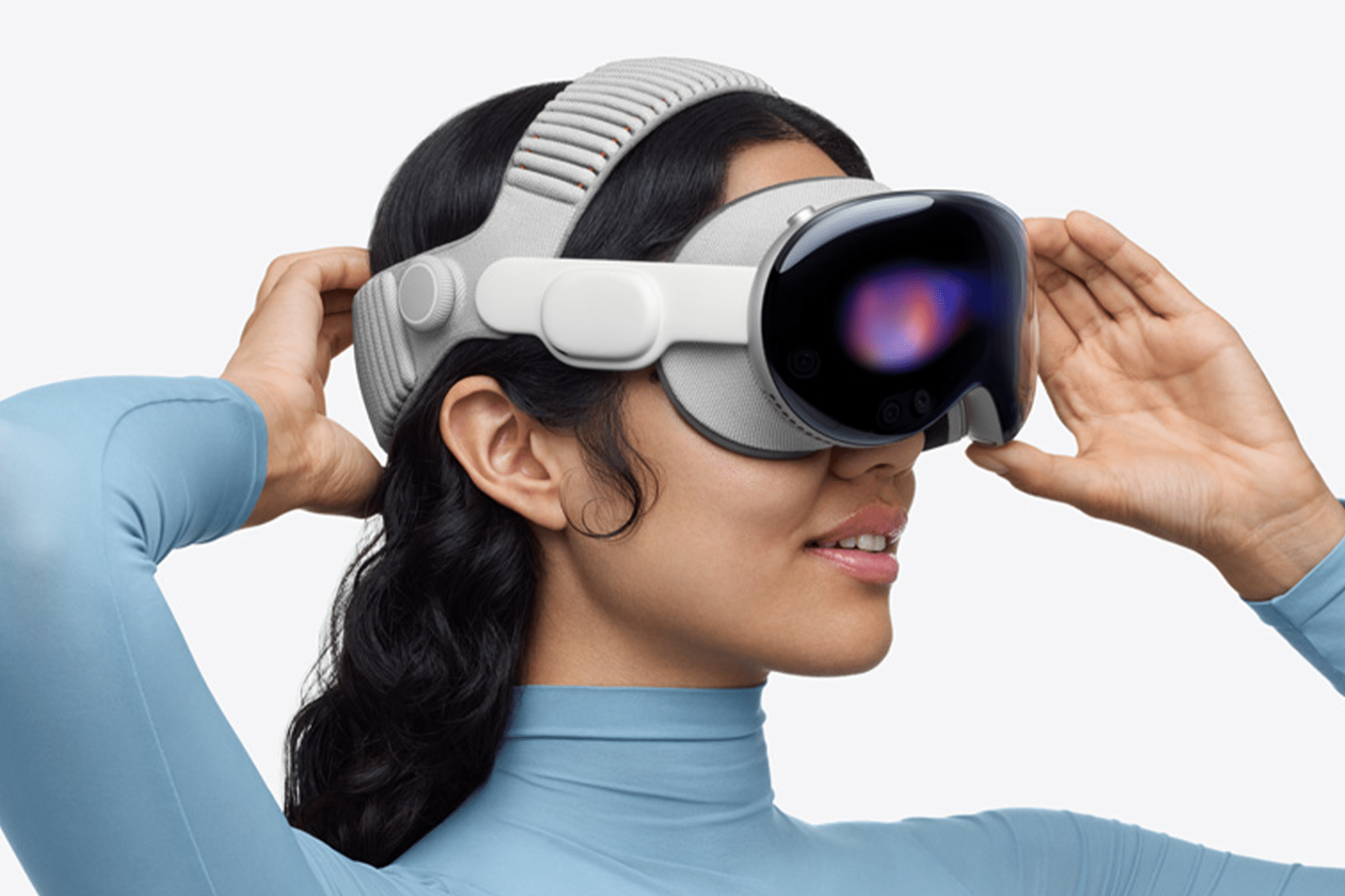A person wearing a light gray virtual reality headset, powered by M5 chip performance, adjusts it with both hands. The individual has long, dark hair and is dressed in a light blue, long-sleeve top.