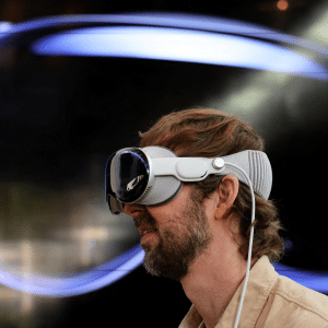 A man with a beard and light brown hair wears what could be the next Apple Vision Pro headset, with a large abstract blue shape displayed on a dark background behind him.