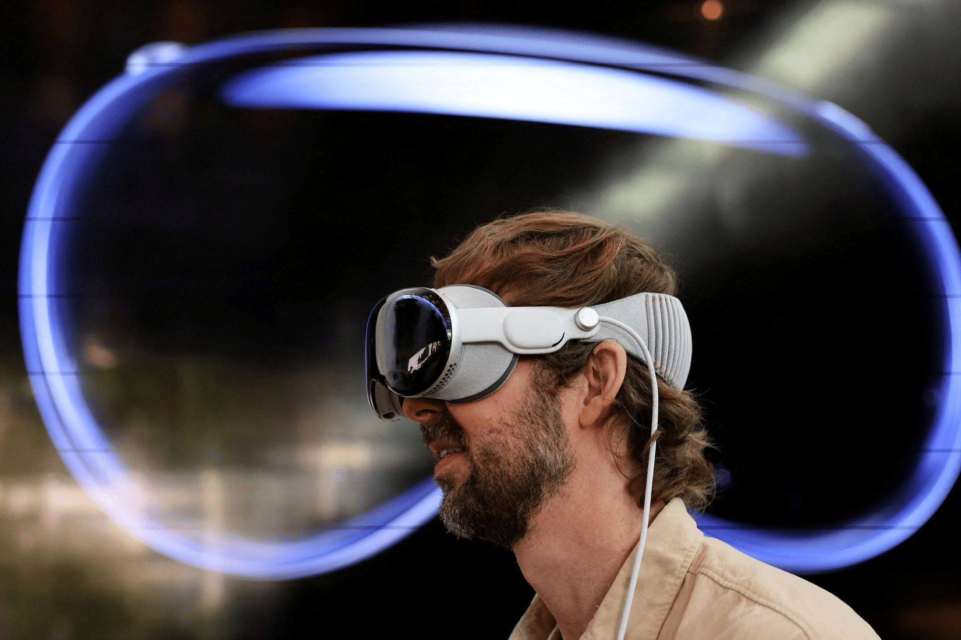 A man with a beard and light brown hair wears what could be the next Apple Vision Pro headset, with a large abstract blue shape displayed on a dark background behind him.