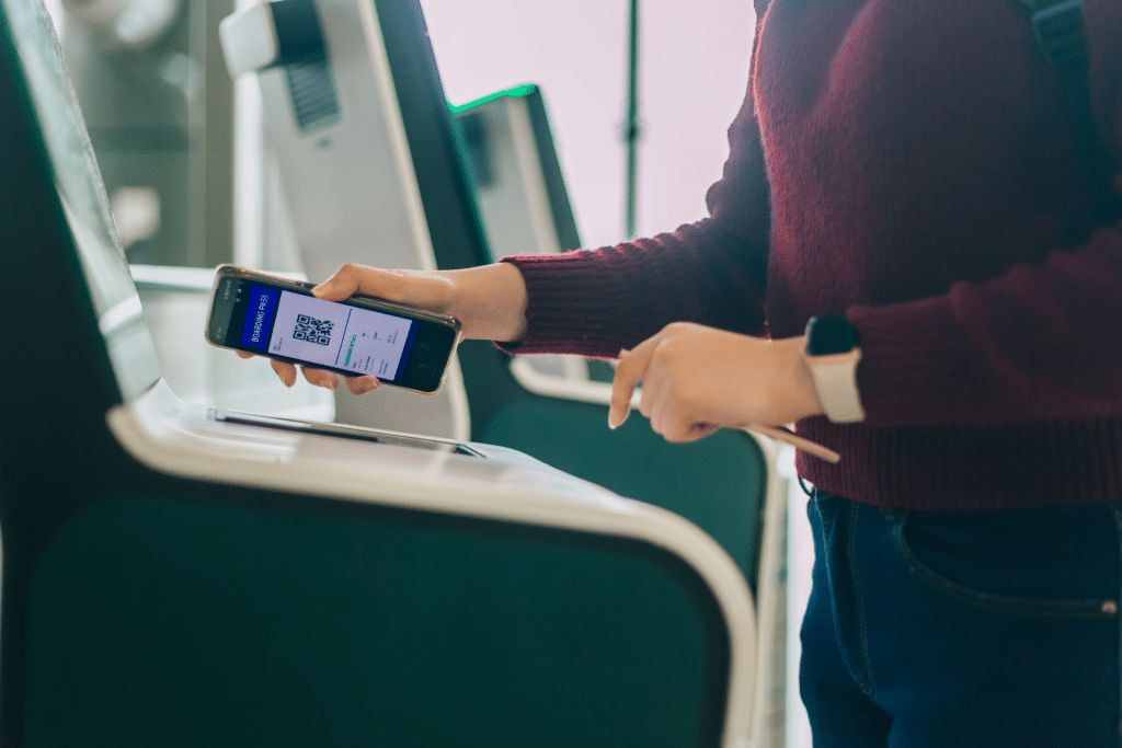 A person scans an Apple Wallet boarding pass from their smartphone at an airport check-in kiosk, wearing a maroon sweater and smartwatch, showcasing the ease of digital travel with iOS 26.