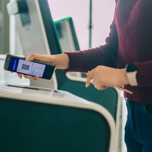 A person scans an Apple Wallet boarding pass from their smartphone at an airport check-in kiosk, wearing a maroon sweater and smartwatch, showcasing the ease of digital travel with iOS 26.