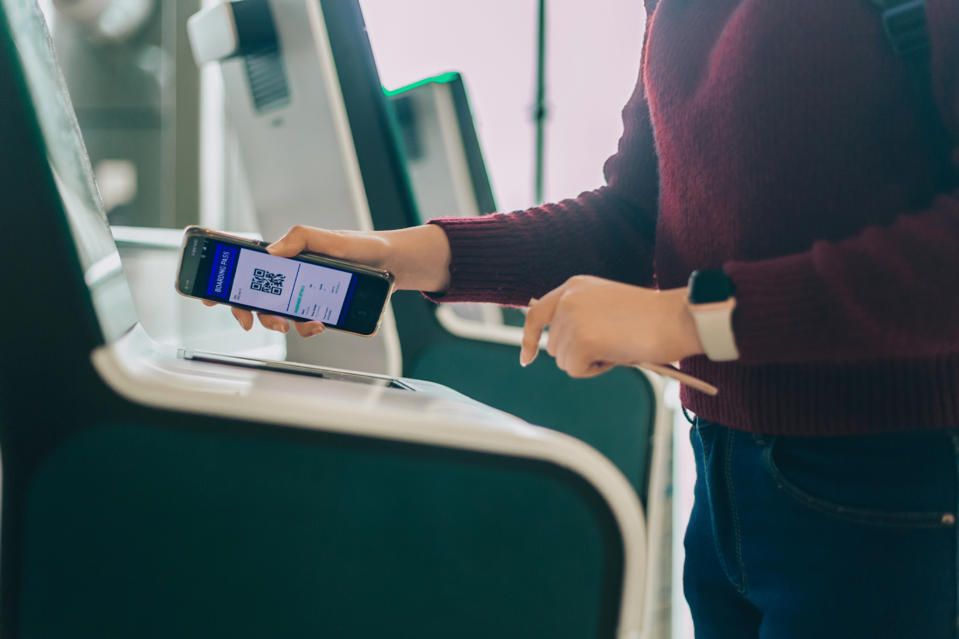 A person scans an Apple Wallet boarding pass from their smartphone at an airport check-in kiosk, wearing a maroon sweater and smartwatch, showcasing the ease of digital travel with iOS 26.