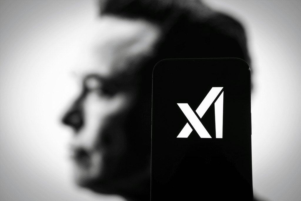 A smartphone displays the white X logo of the X social media app, hinting at xAI and AI-powered video game integration, with the blurred side profile of a man's face in the background.