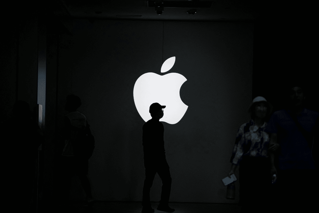 A person stands silhouetted in front of a large, illuminated Apple logo on a dark wall—perhaps reflecting on the growth of the Apple Services division—as others move through the dimly lit space, highlighting the dramatic contrast.