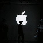 A person stands silhouetted in front of a large, illuminated Apple logo on a dark wall—perhaps reflecting on the growth of the Apple Services division—as others move through the dimly lit space, highlighting the dramatic contrast.