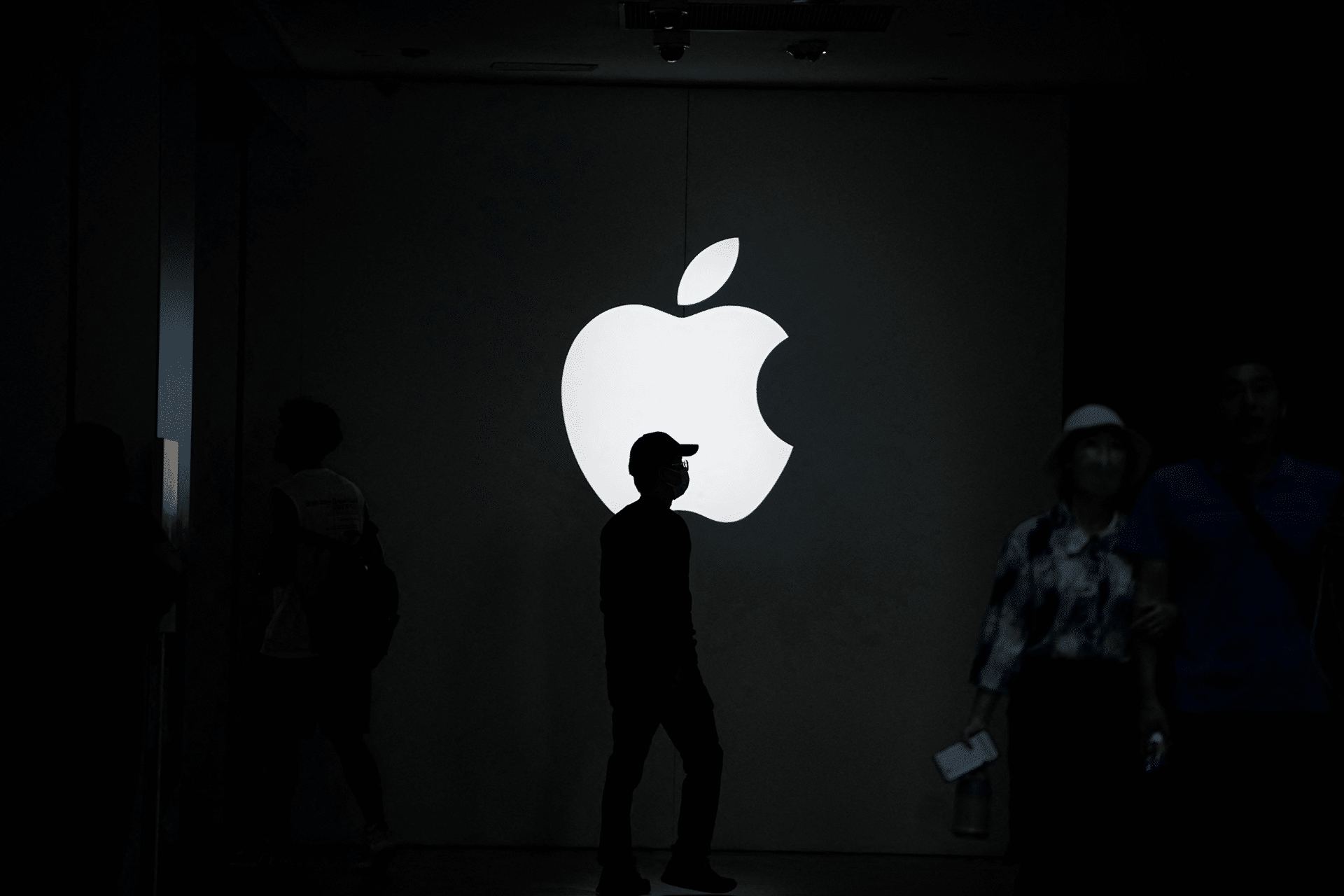 A person stands silhouetted in front of a large, illuminated Apple logo on a dark wall—perhaps reflecting on the growth of the Apple Services division—as others move through the dimly lit space, highlighting the dramatic contrast.