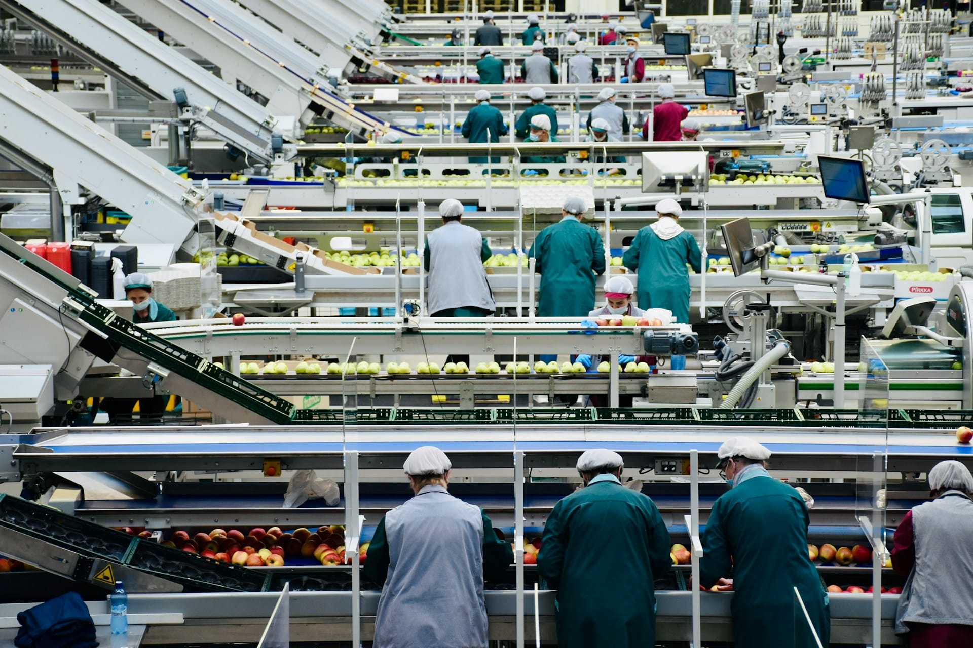 Workers in hairnets and uniforms sort apples on conveyor belts in a large, modern fruit processing facility with automated machinery, computer screens, and top-tier food plant sanitation services ensuring cleanliness throughout.