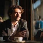 A man with curly hair and a suit jacket sits at a café table by a sunlit window, holding a smartphone and smiling thoughtfully. A coffee cup rests in front of him, and blurred figures are visible outside.