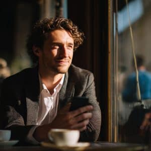 A man with curly hair and a suit jacket sits at a café table by a sunlit window, holding a smartphone and smiling thoughtfully. A coffee cup rests in front of him, and blurred figures are visible outside.