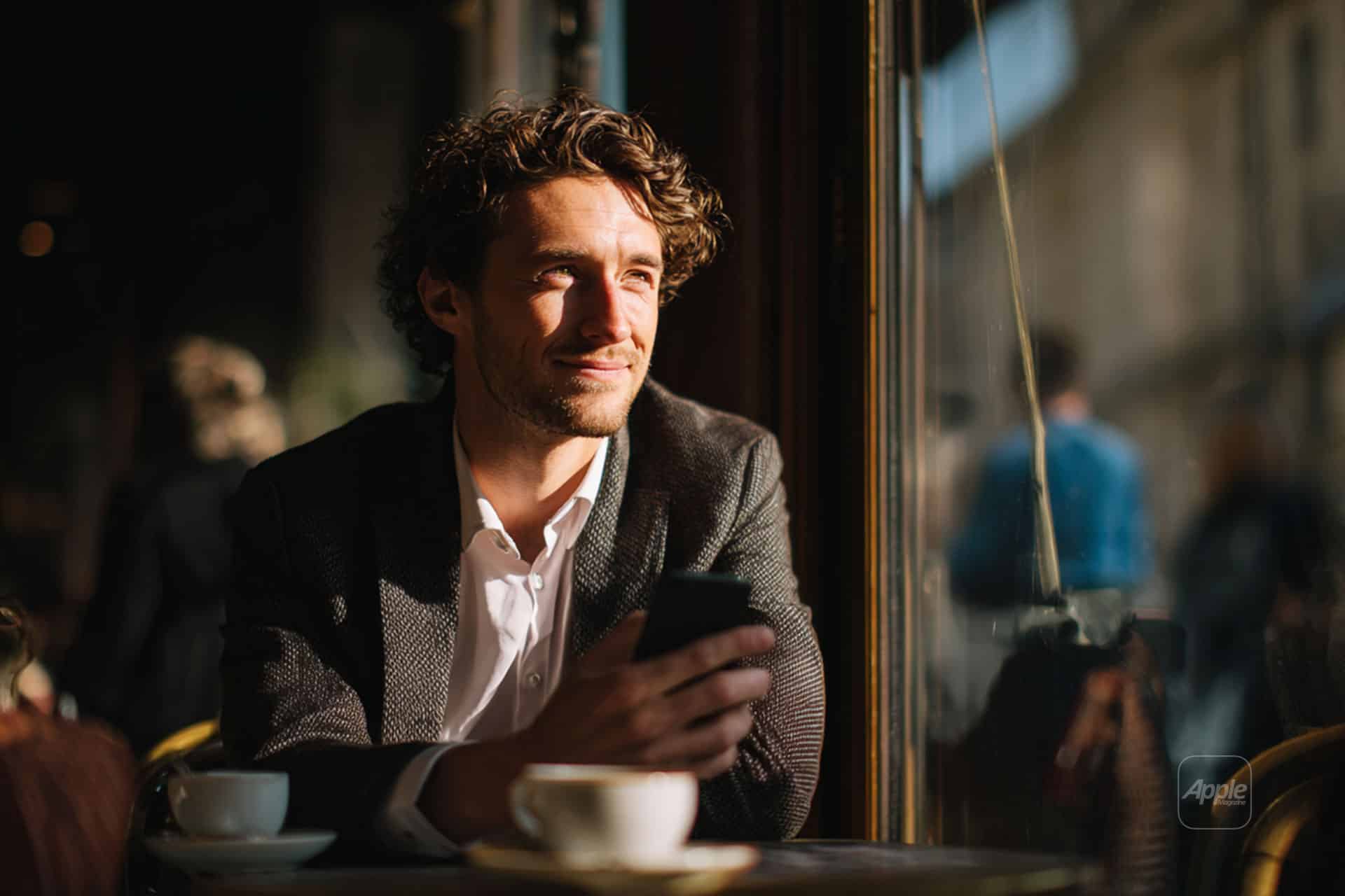 A man with curly hair and a suit jacket sits at a café table by a sunlit window, holding a smartphone and smiling thoughtfully. A coffee cup rests in front of him, and blurred figures are visible outside.