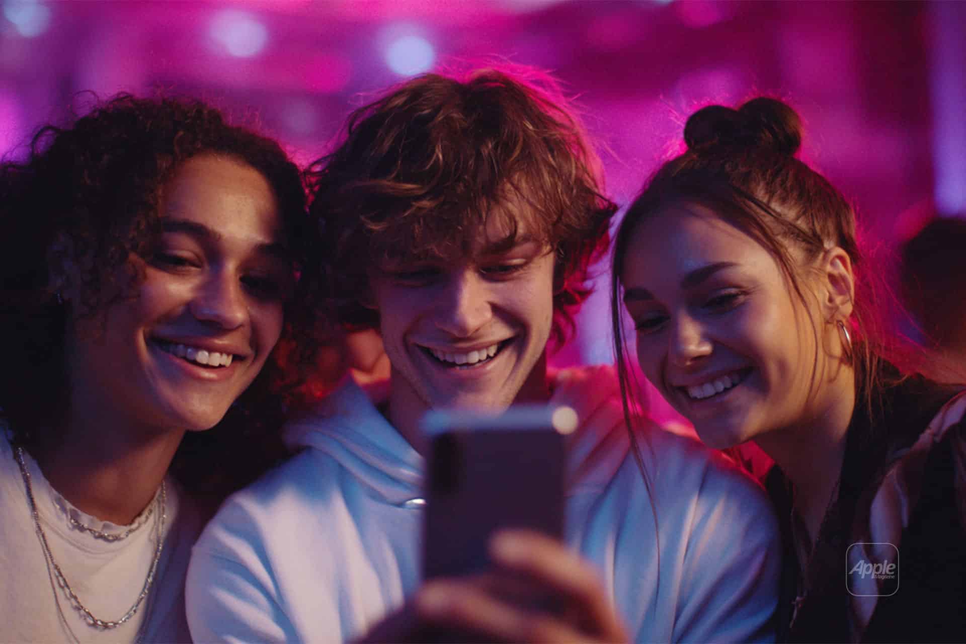 Three smiling young people take a selfie together in a vibrant, pink-lit setting. They look happy and close, with their faces illuminated by the phone’s screen.