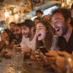 A group of young adults sit at a bar, laughing and cheering while looking at their phones. There are drinks on the wooden counter and a lively, warm atmosphere in the background.
