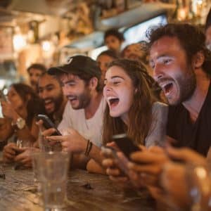 A group of young adults sit at a bar, laughing and cheering while looking at their phones. There are drinks on the wooden counter and a lively, warm atmosphere in the background.