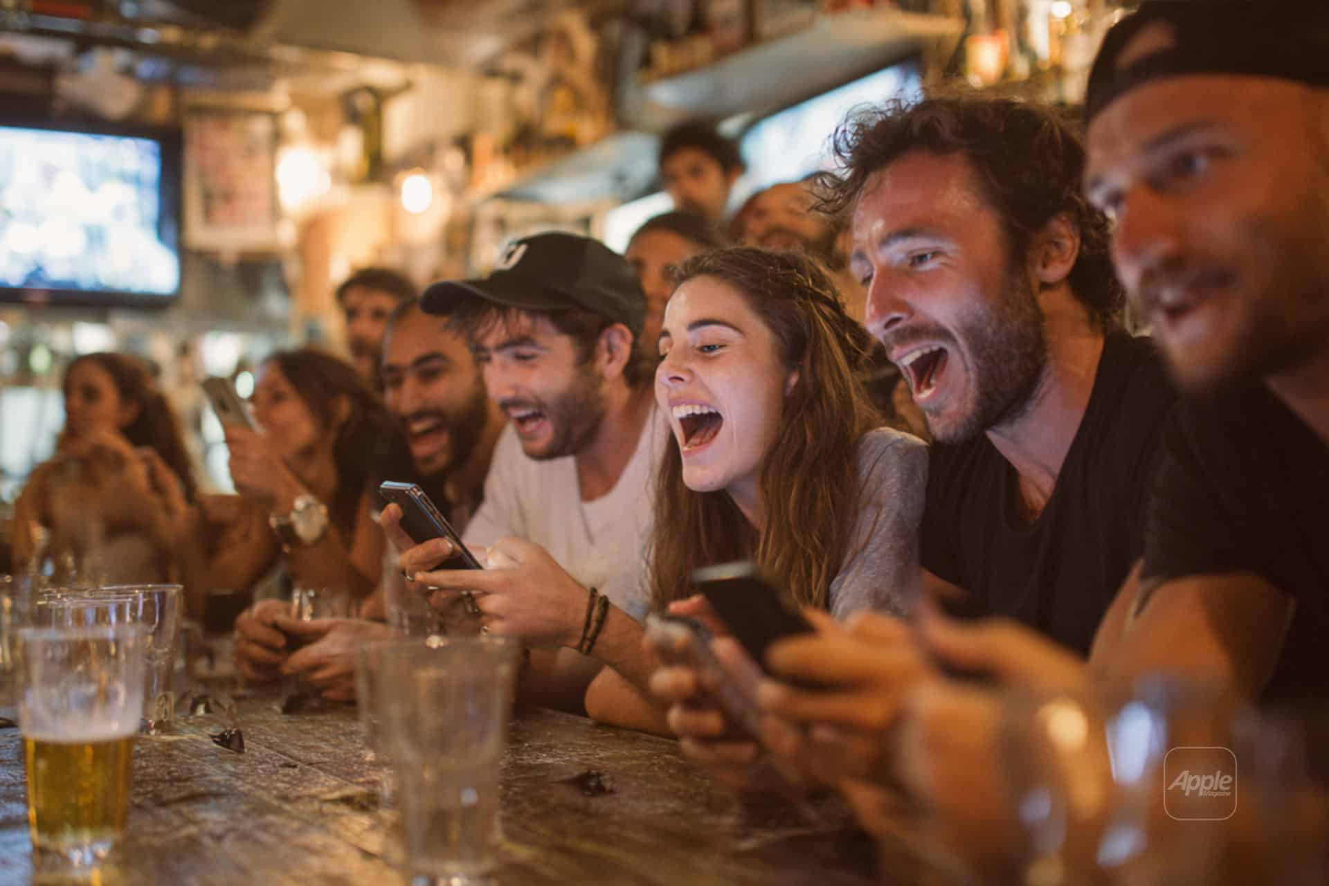 A group of young adults sit at a bar, laughing and cheering while looking at their phones. There are drinks on the wooden counter and a lively, warm atmosphere in the background.