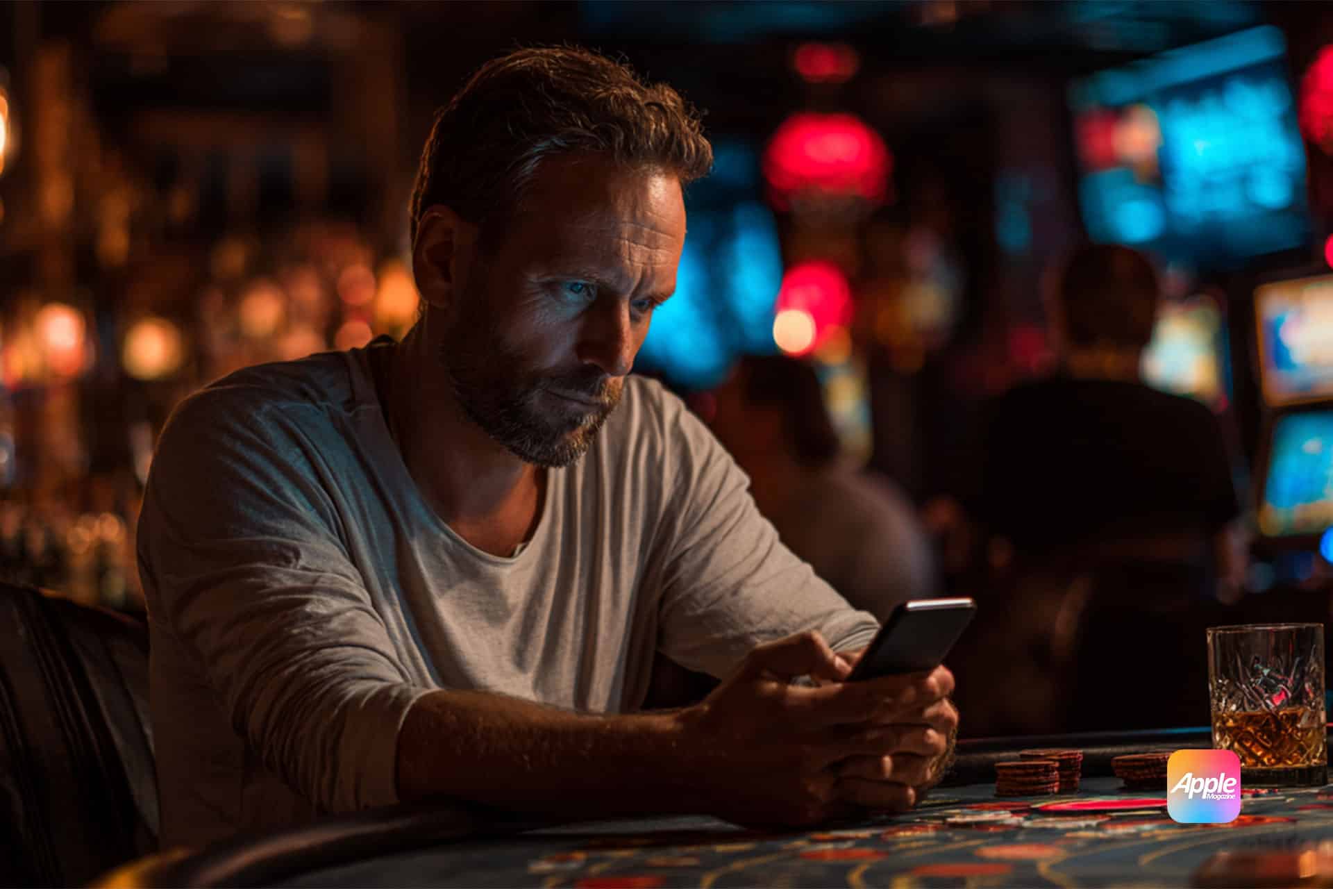 A man sits at a casino table, focused on his smartphone. Poker chips and a drink are nearby. The dimly lit background shows casino decor and other people. An Apple logo is visible on the table.