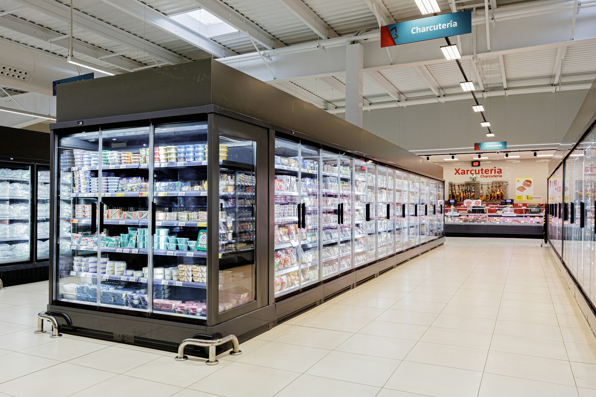 A supermarket aisle with glass-door refrigerated displays filled with dairy products and packaged foods; a sign above reads "Charcutería" near the deli section, where regular food plant sanitation ensures cleanliness and safety.