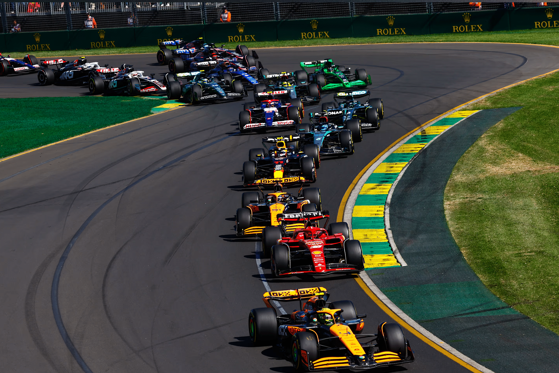 A group of Formula 1 cars race closely together on a curving track, surrounded by green grass and safety barriers, as drivers navigate a sharp turn in a scene reminiscent of F1 The Movie during a competitive event.