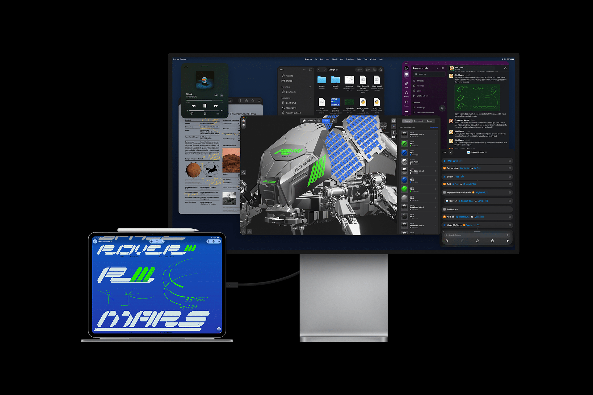 A desktop monitor displays various open applications and a robotic model. In front, an M5 lineup iPad showcases futuristic blue and green text graphics, connected to the monitor, suggesting a multitasking workspace setup and highlighting iPad innovation.