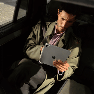 A man wearing a trench coat sits in the backseat of a car, looking intently at his iPad Pro with the M5 chip, using it with one hand. Sunlight partially illuminates him as he works.